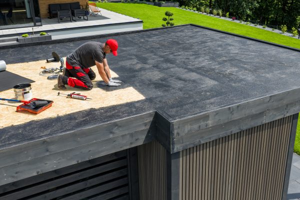A construction worker applies EPDM roofing material on a flat roof of a modern home in a sunny outdoor environment. Landscaping is visible in the background.